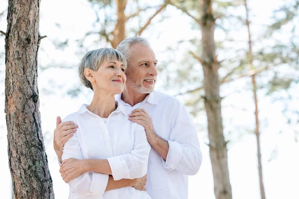 Smiling Senior Couple White Shirts Embracing Looking Away Forest — Stock Photo, Image