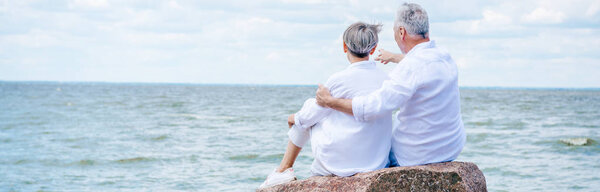 panoramic view of senior couple sitting on stone and embracing near river