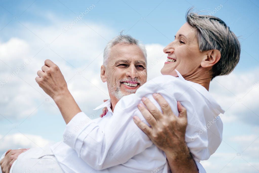 smiling senior man in white shirt holding wife under blue sky