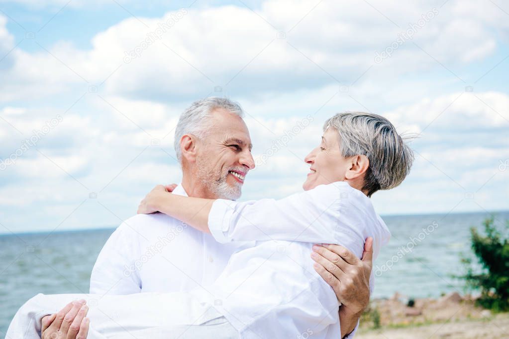 smiling senior man in white shirt lifting wife under blue sky