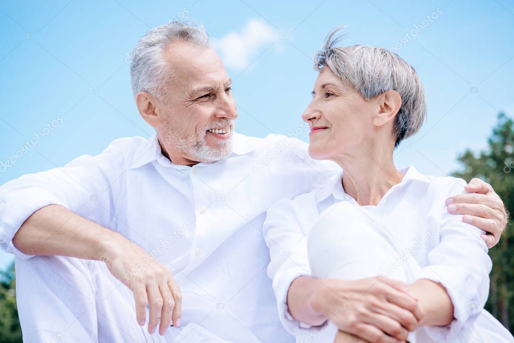 happy smiling senior couple in white shirts embracing and looking at each other under blue sky