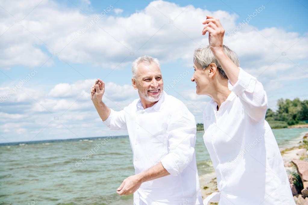 happy smiling senior couple in white shirts looking at each other and gesturing at beach under blue sky