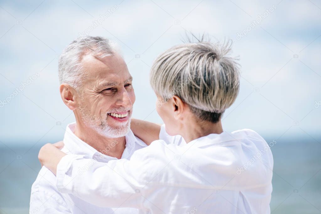 happy senior couple in white shirts embracing and looking at each other under blue sky