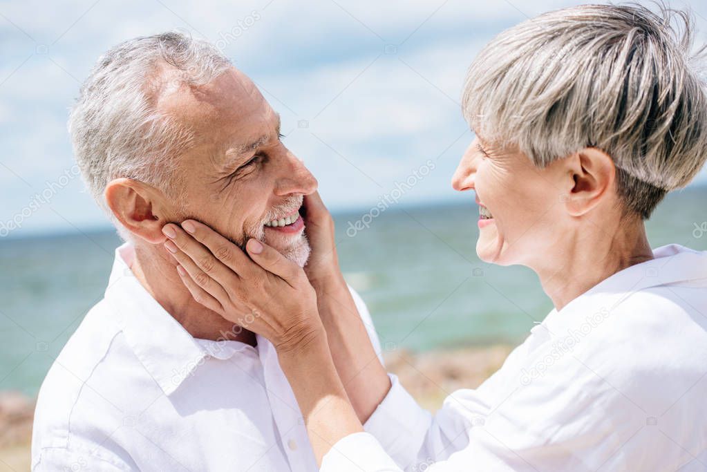 happy senior woman looking at husband and touching his face under blue sky