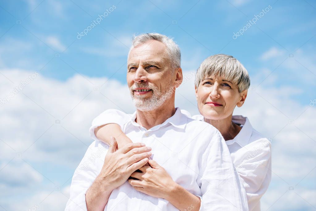 happy senior couple in white shirts embracing under blue sky