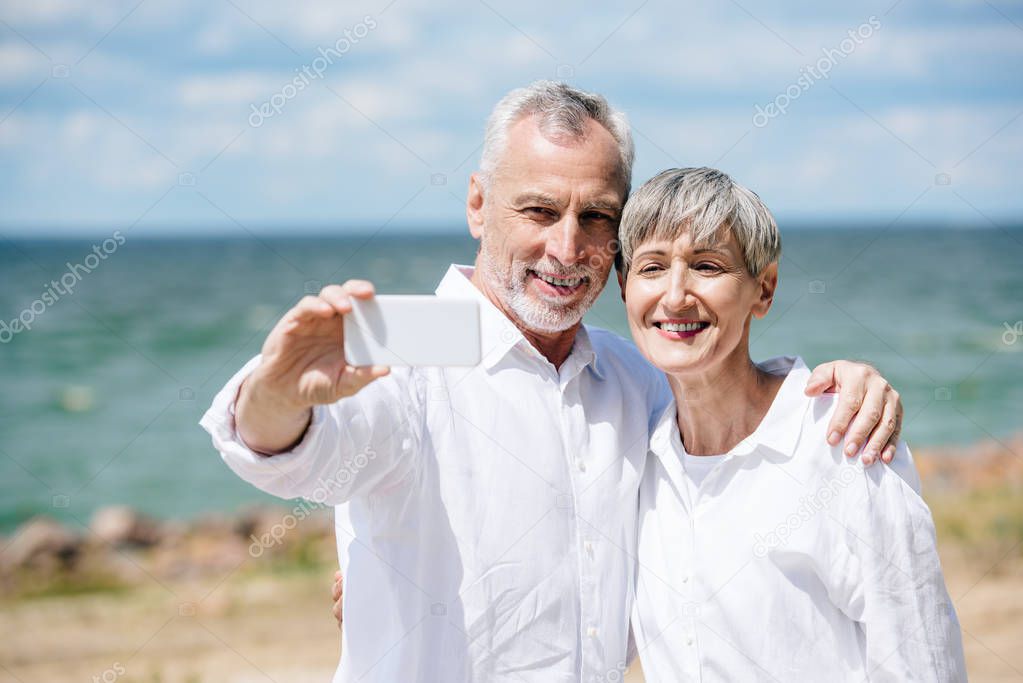 smiling senior couple in white shirts embracing and taking selfie at beach