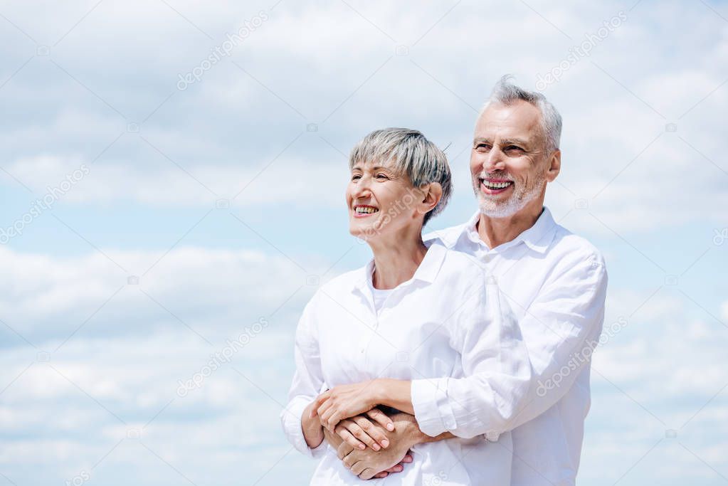 happy senior couple in white shirts embracing under blue sky