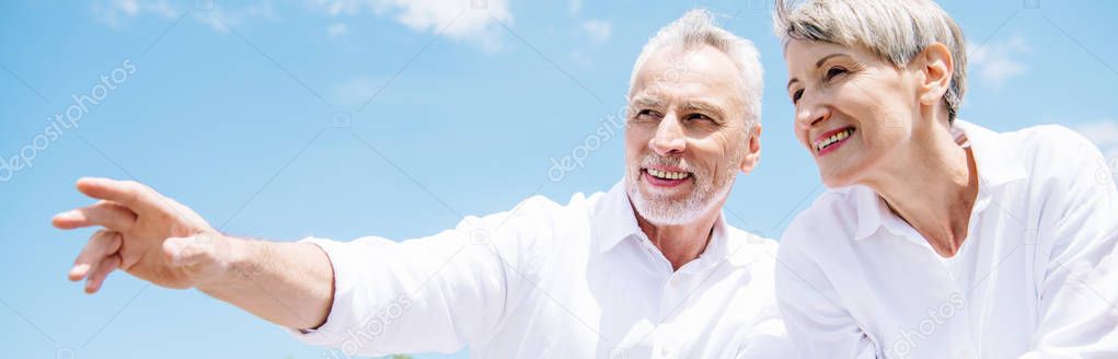 panoramic view of smiling senior couple in white shirts looking away under blue sky