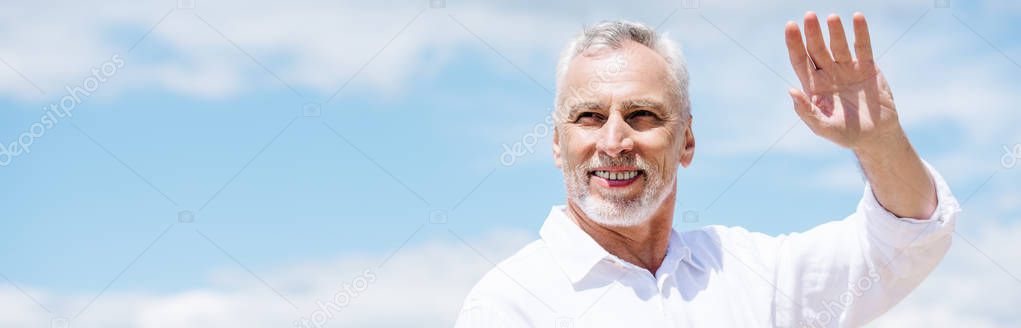 panoramic view of smiling senior man in white shirt waving hand under blue sky