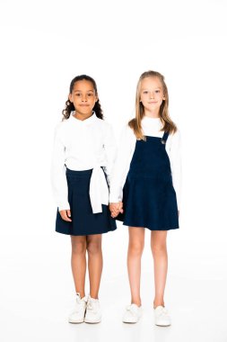 full length view of two multicultural schoolgirls holding hands on white background
