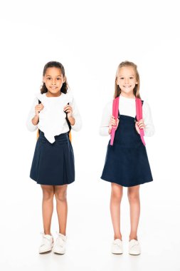 full length view of two adorable multicultural schoolgirls smiling at camera on white background