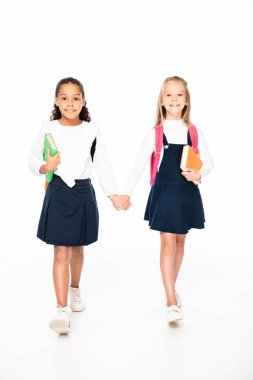 full length view of two smiling multicultural schoolgirls walking while holding hands on white background