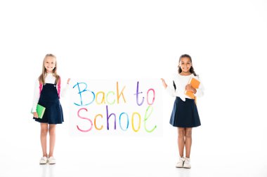 full length view of two multicultural schoolgirls holding books and placard with back to school inscription on white background