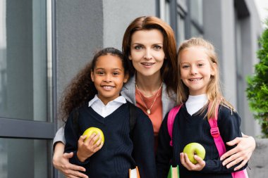 happy mother hugging cheerful multicultural daughters and smiling at camera while standing near school  clipart