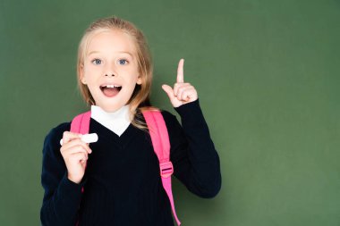 cheerful schoolgirl holding piece of chalk and showing idea gesture while standing near green chalkboard clipart