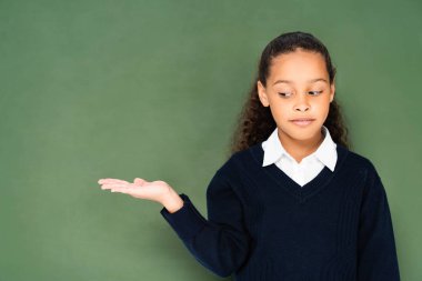 thoughtful, skeptical schoolgirl pointing with hand at green chalkboard  clipart