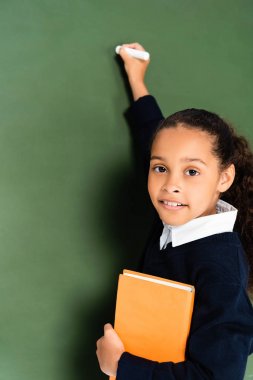 kitap tutan ve kamera bakarken tebeşir tahtası üzerinde yazma sevimli afro-amerikan kız öğrenci