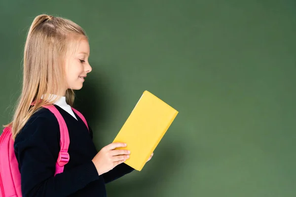 緑の黒板の近くに立ちながら本を読む笑顔の女子高生の側面図 — ストック写真