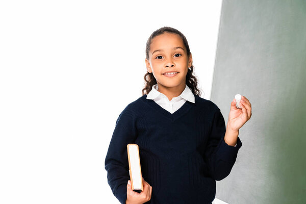 прелестная африканская школьница, стоящая рядом с доской с книгой и мелом, изолированным на белом
