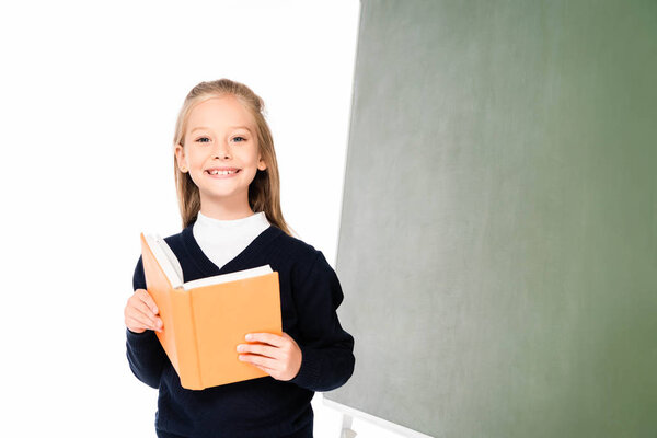 adorable schoolgirl smiling at camera while standing near chalkboard and holding book isolated on white