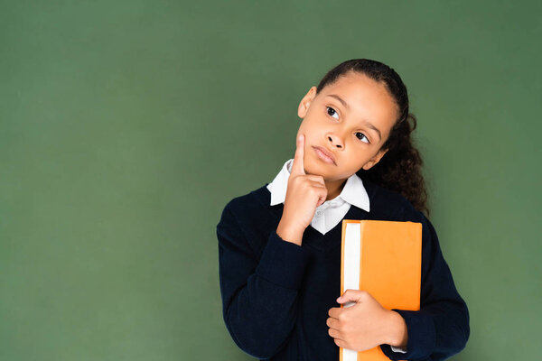 вдумчивая африканская школьница держит книгу и смотрит в сторону, стоя рядом с доской