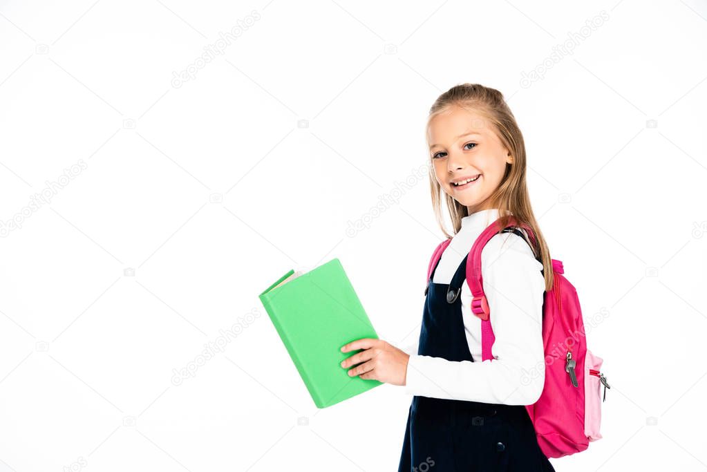 adorable schoolgirl holding book and smiling at camera isolated on white