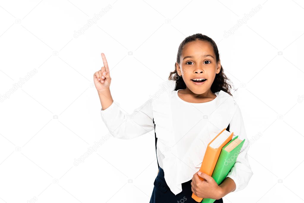 cheerful african american schoolgirl pointing with finger while holding books isolated on white