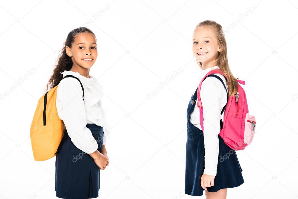 two cheerful multicultural schoolgirls with backpacks looking at camera isolated on white