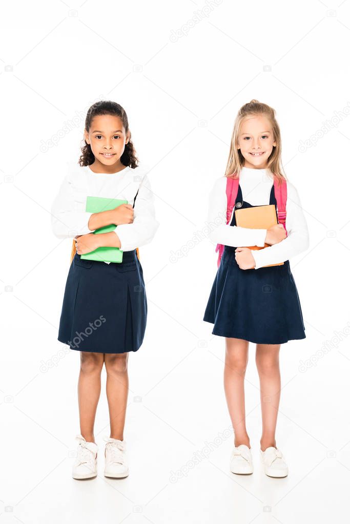 full length view of two adorable multicultural schoolgirls holding books on white background