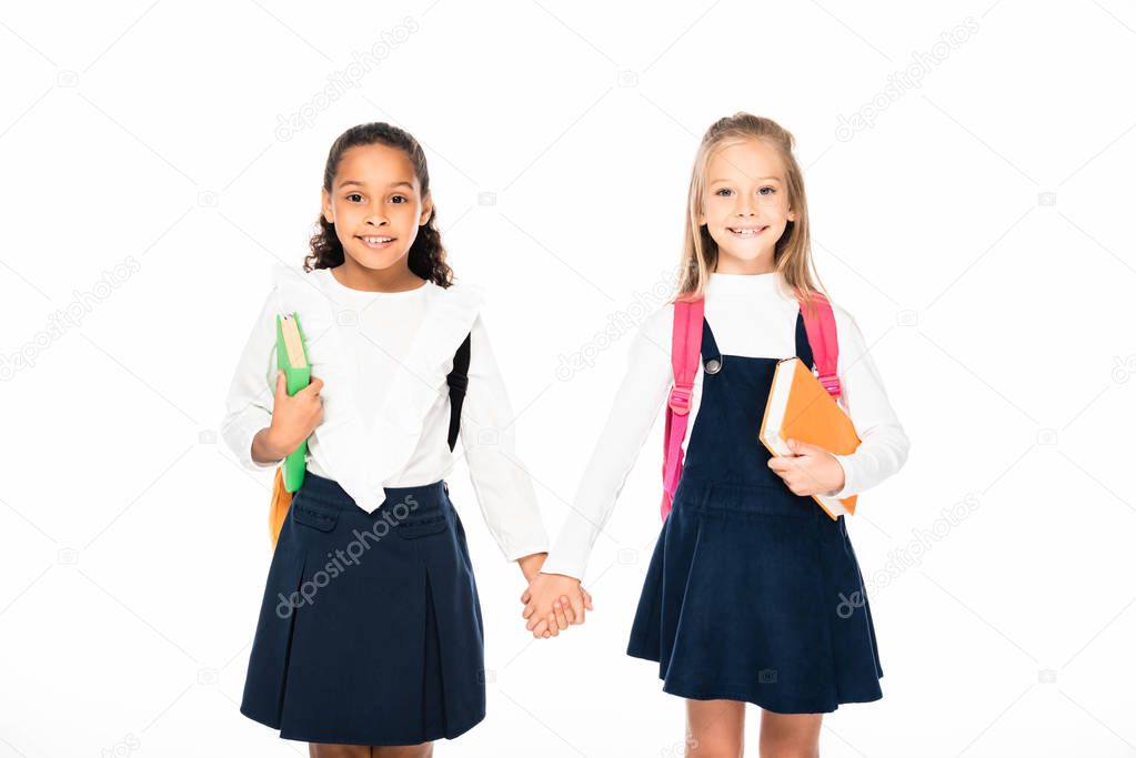 two adorable multicultural schoolgirls holding hands and looking at camera isolated on white