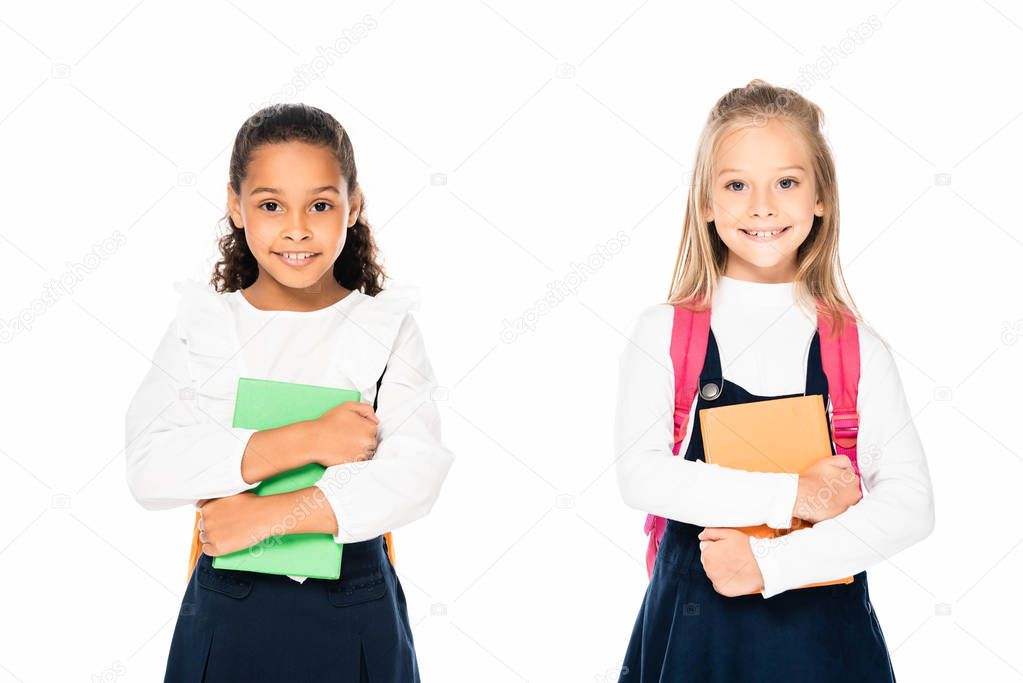 two cute multicultural schoolgirls holding books and smiling at camera isolated on white