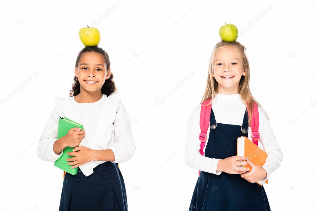 two cheerful multicultural schoolgirls with apples on heads holding books isolated on white
