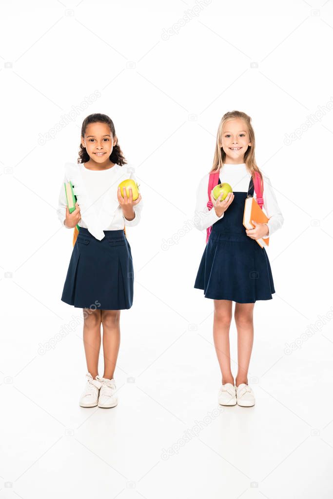 full length view of two smiling multicultural schoolgirls holding books and apples on white background