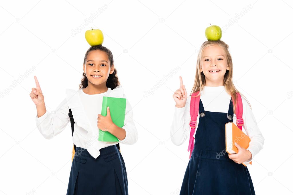 two cheerful multicultural schoolgirls with apples on heads showing idea gestures isolated on white