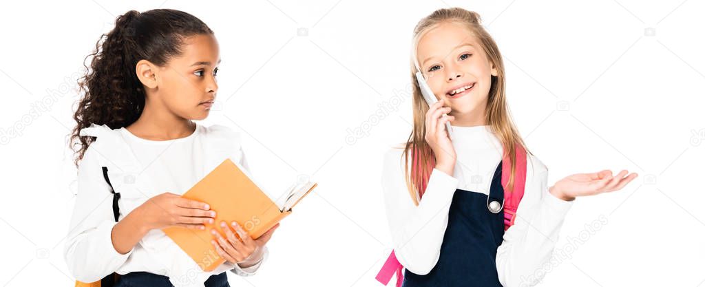 panoramic shot of displeased african american schoolgirl looking at cheerful friend talking on smartphone isolated on white