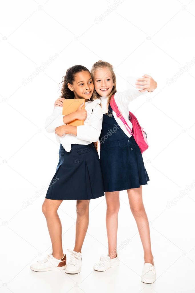 full length view of happy schoolgirl hugging african american friend and taking selfie on white background
