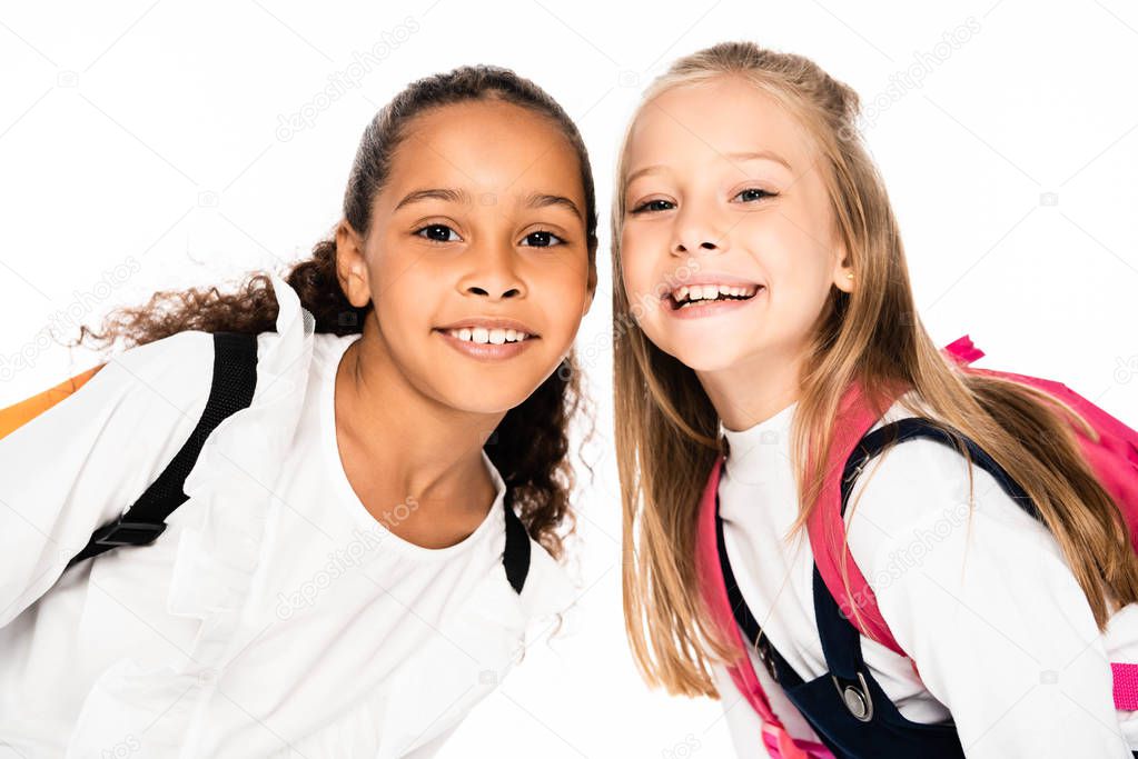 two adorable multicultural schoolgirls smiling while looking at camera isolated on white