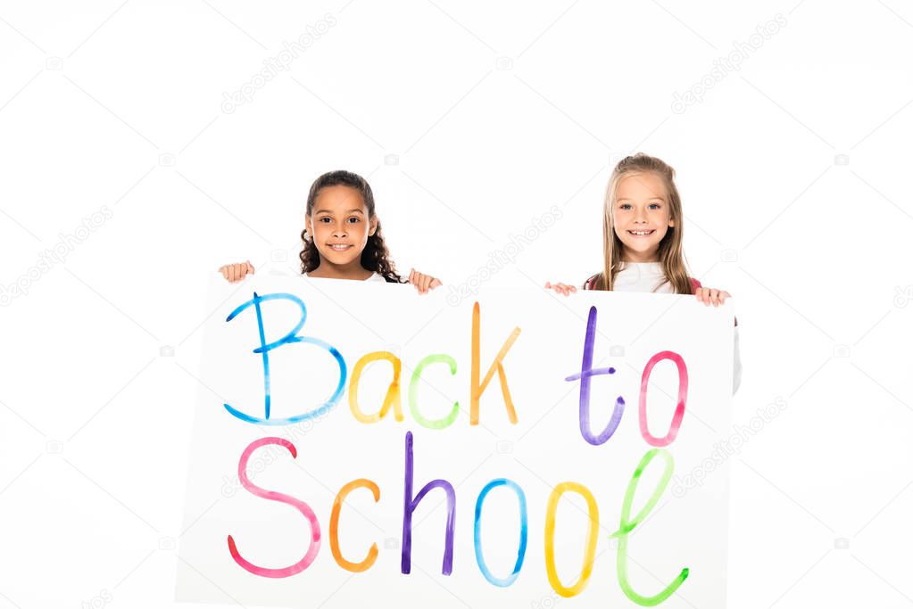 two adorable multicultural schoolgirls holding placard with back to school inscription isolated on white