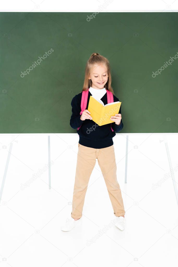full length view of schoolgirl reading book while standing near green chalkboard on white background