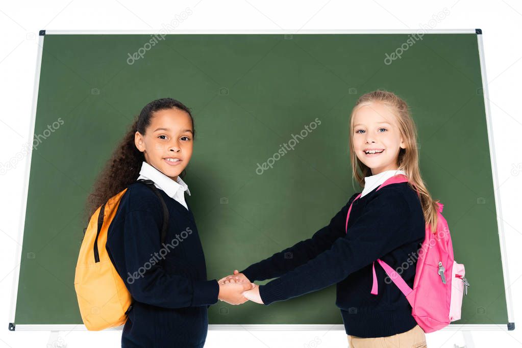 two adorable multicultural schoolgirls holding hands while standing near chalkboard isolated on white