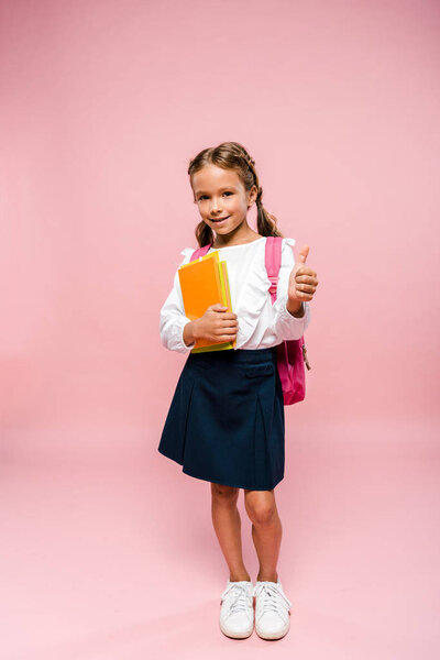 happy kid holding books while standing with backpack and showing thumb up on pink 