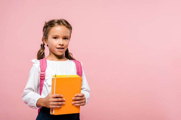 happy kid holding books while standing with backpack isolated on pink 