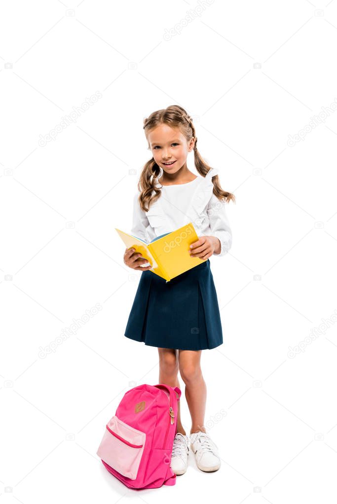 happy child reading book and standing near pink backpack isolated on white 