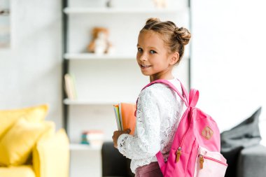 sırt çantası ile ayakta iken kitap tutan gülümseyen okul çocuğu 