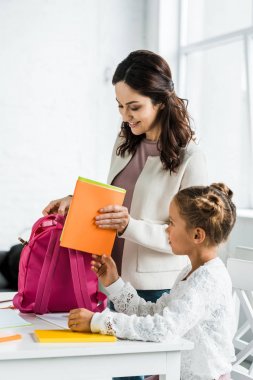 mutlu anne evde kız öğrencinin sırt çantasında kitap koyarak 