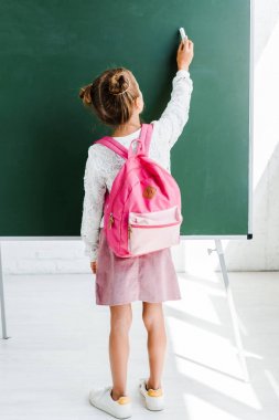 sırt çantası ile ayakta ve yeşil tebeşir tahta yakınında tebeşir tutan kız öğrencinin arka görünümü 