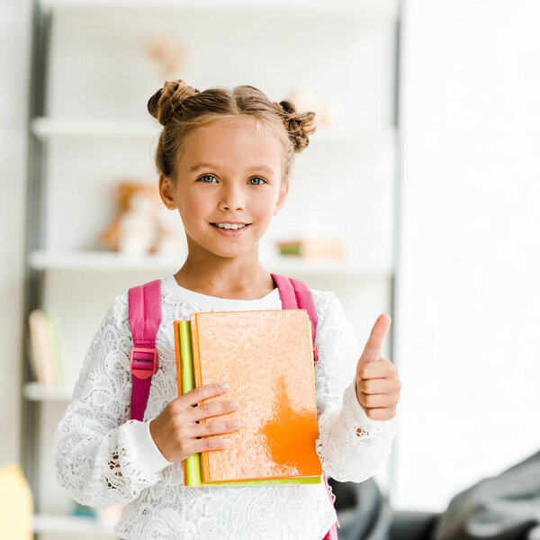 cheerful schoolgirl holding books and showing thumb up at home