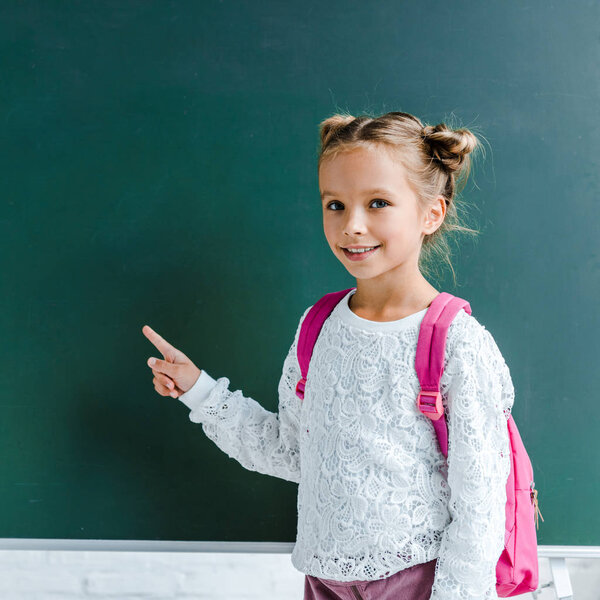 happy kid smiling while pointing with finger at green chalkboard 