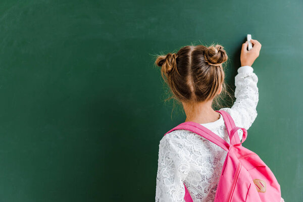 back view of schoolchild holding chalk near chalkboard on green 