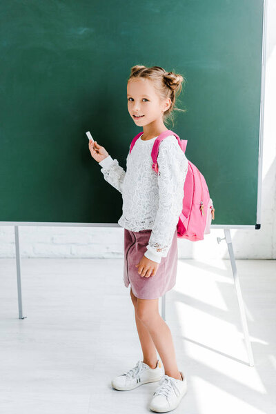 cute schoolgirl standing with backpack and holding chalk near green chalkboard 